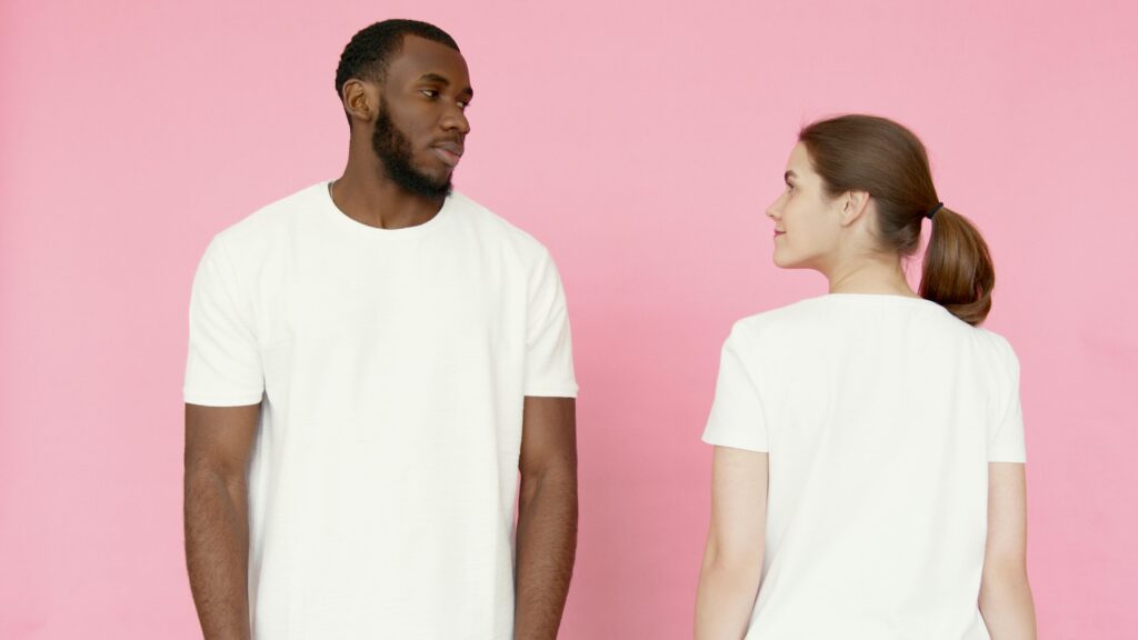 A man and woman in white t-shirts stand against a pink backdrop.