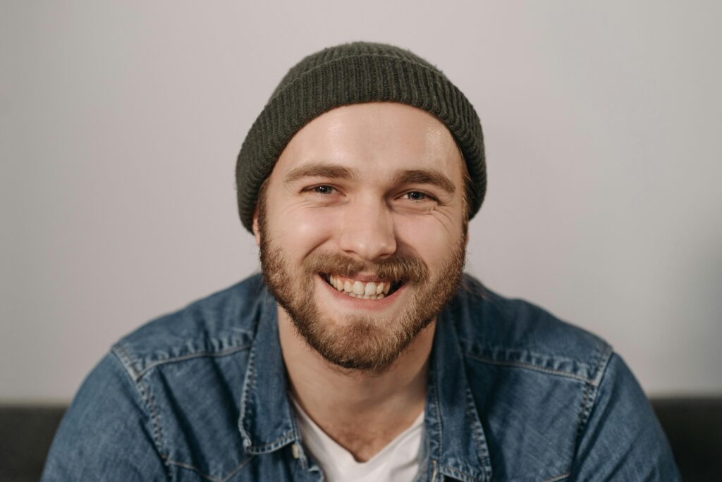 Close-up portrait of a cheerful young man with a beard and knit cap wearing denim.
