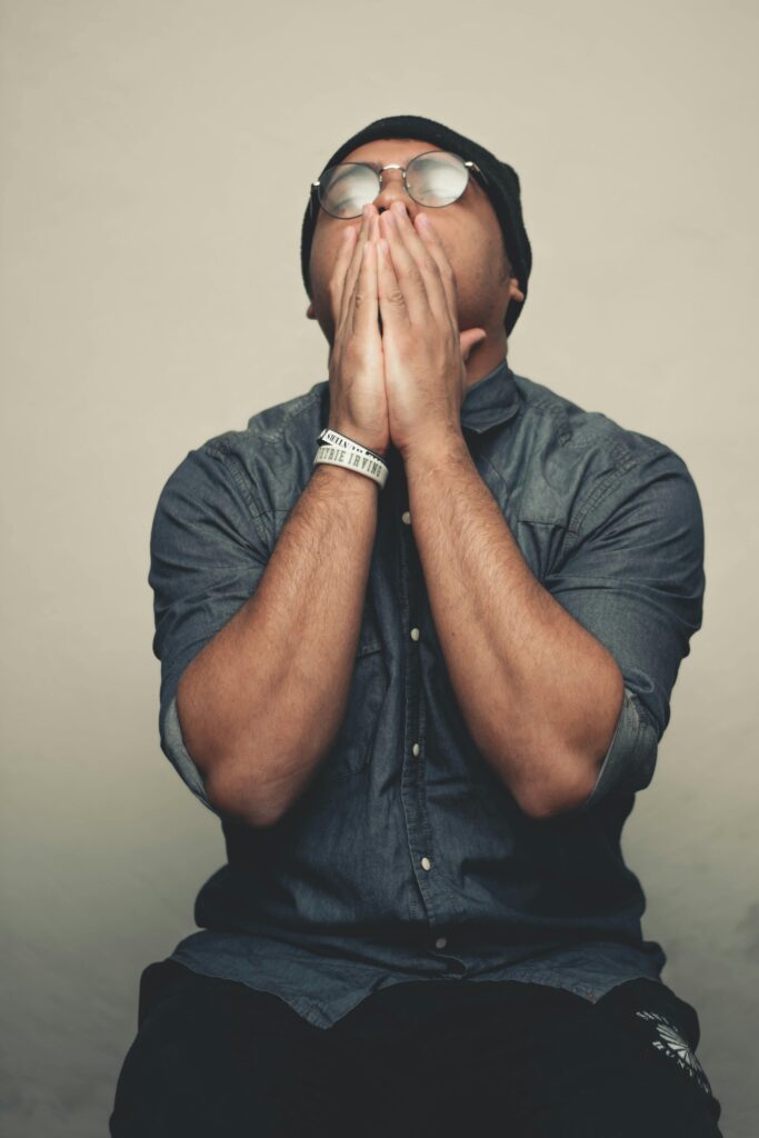 Portrait of a young man indoors, displaying stress with hands on face, wearing casual outfit.