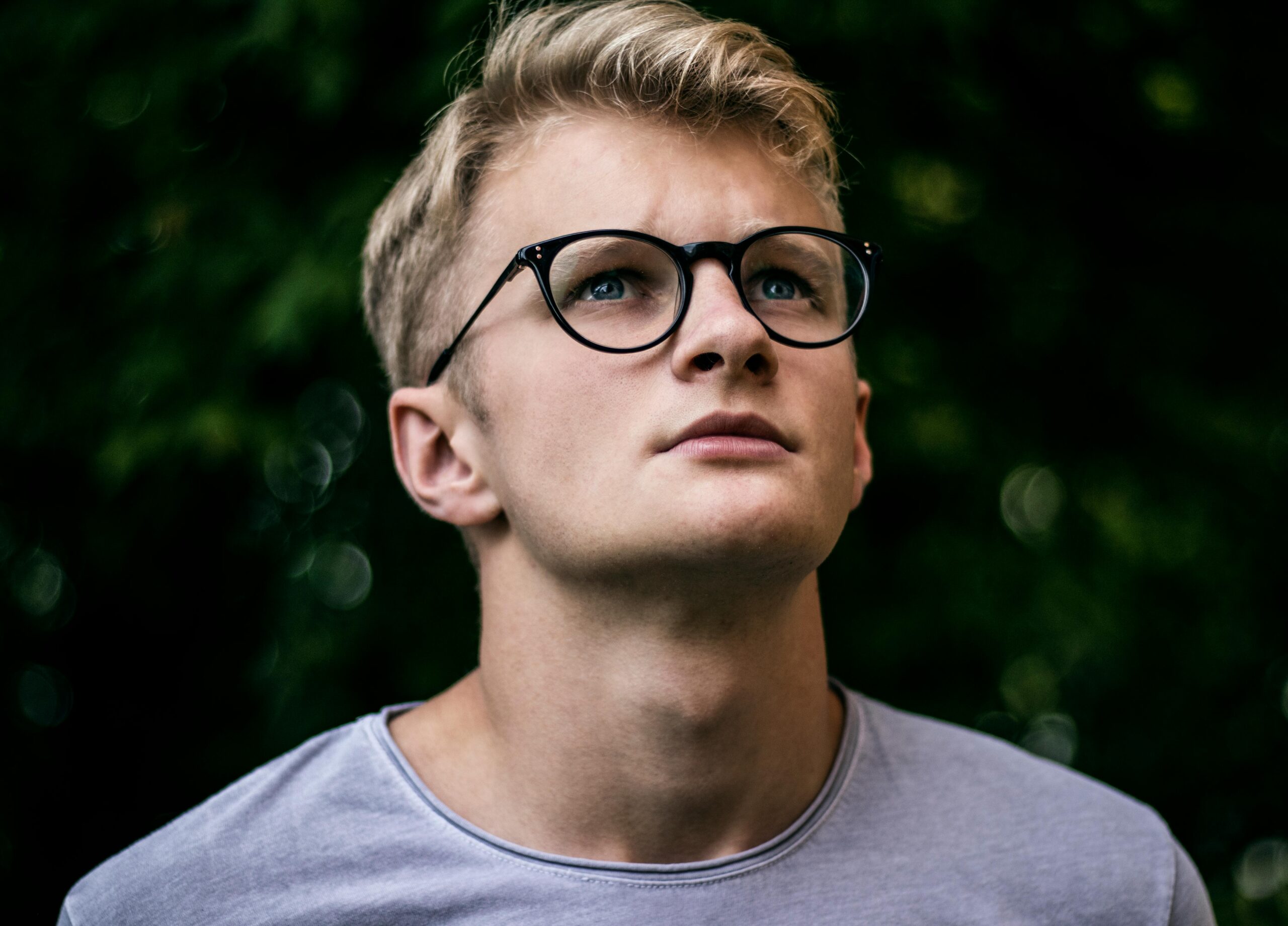 A thoughtful young man wearing eyeglasses looks upwards in an outdoor setting.