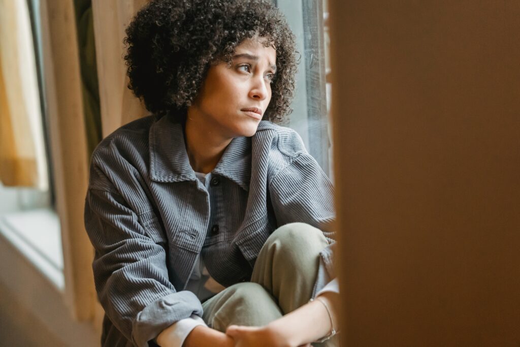 Frowning upset African American female with curly hair embracing knees while sitting on windowsill